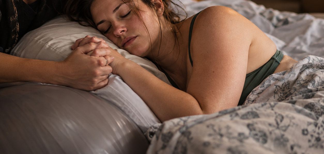 A laboring woman rests on a pillow with her eyes closed, holding her doula's hands.