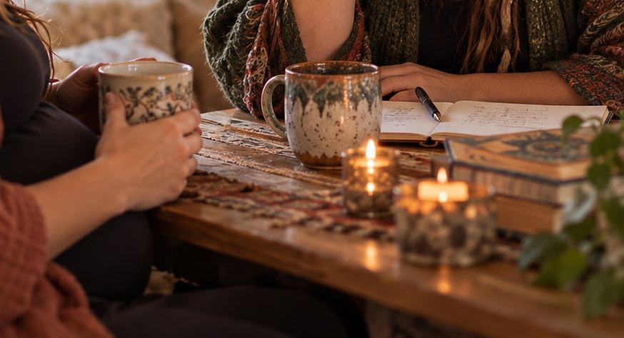 Two mugs, a notebook and lit candles on a low table between two people talking.